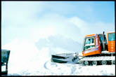 Фото оранжевого ратрака на переднем плане, вдали - горы в снежной дымке